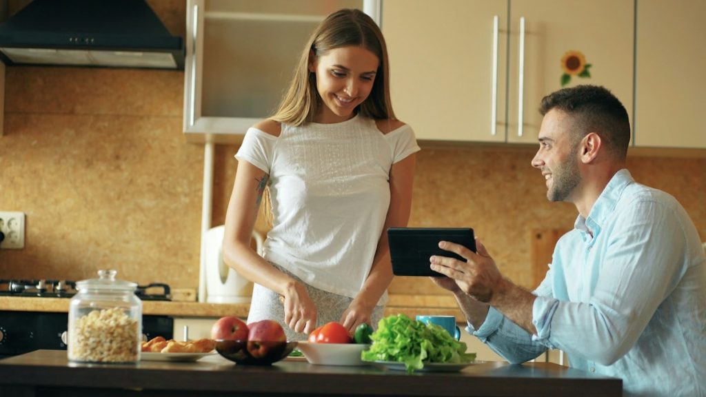 🎬 FULL 4K VIDEO for this still is in my bio link! ⬇️ Attractive couple chatting in the kitchen early morning. Handsome man using tablet while his girlfriend cooking breakfast at home